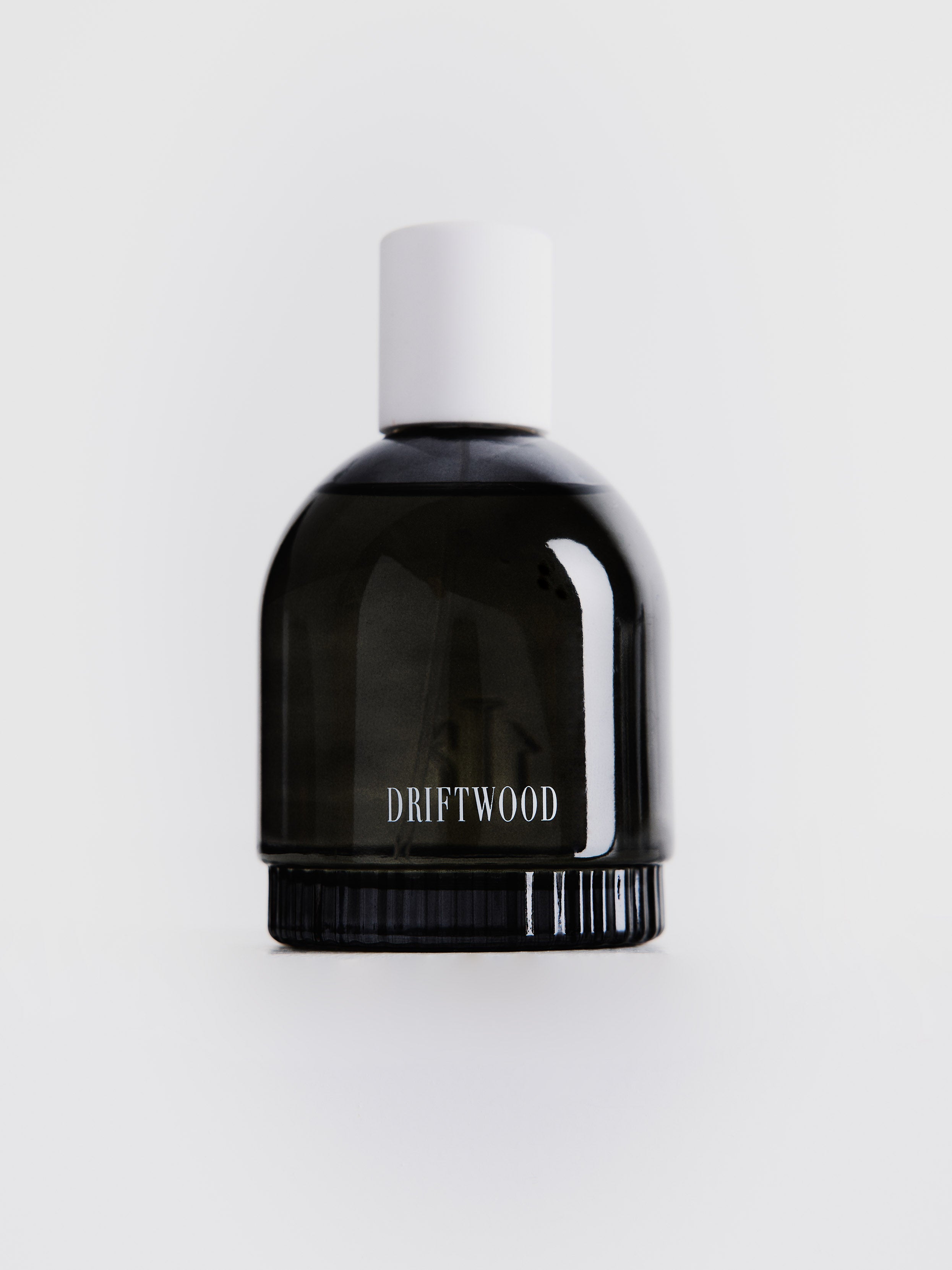 Small dark gray transparent perfume bottle with white "DRIFTWOOD" label and white cup on top 